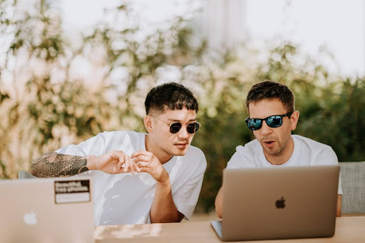 Two men in sunglasses at a table with a laptop, engaged in a discussion.